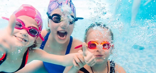 drei Kinder mit Badekappe und Schwimmbrille unter Wasser lachen in die Kamera